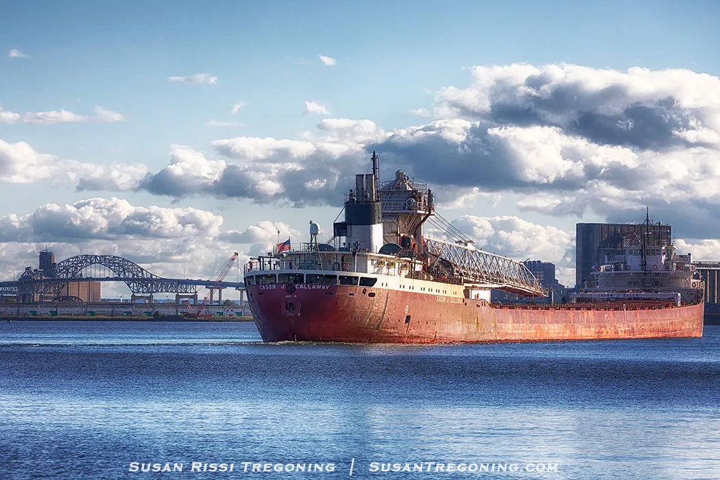 The freighter Cason J. Callaway moves through the Duluth Harbor with the Blatnik Bridge visible in the background. The ship’s red and white hull and deck equipment are brightly lit by the sun, and industrial buildings and cranes line the shoreline under a partly cloudy sky.