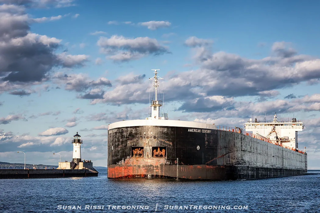 
The Great Lakes freighter American Century approaches the Duluth Ship Canal on a partly cloudy day, passing the North Pier Light on the left. The ship’s dark hull shows areas of rust near the waterline, and the lighthouse stands on a concrete pier extending into the lake. The water is calm and reflects the ship and sky.