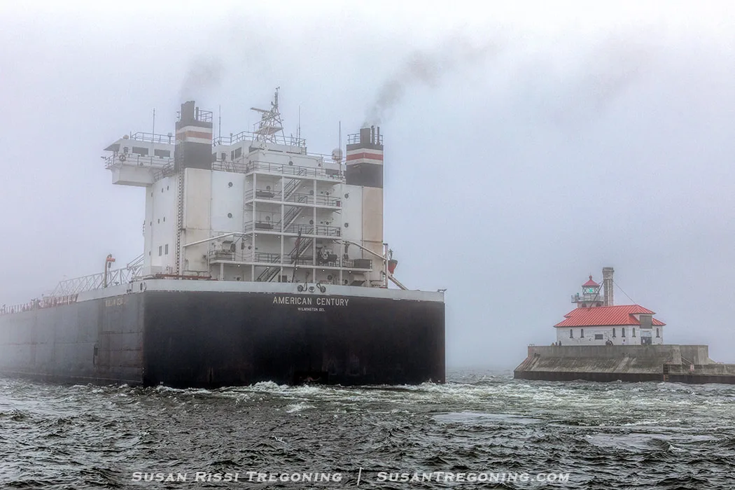 A large cargo ship, the American Century, moves through fog on Lake Superior as it passes the South Breakwater Outer Light at the end of the Duluth Ship Canal. The ship emits dark exhaust, and the lighthouse stands on a concrete pier to the right. The water is choppy under the gray, misty sky.