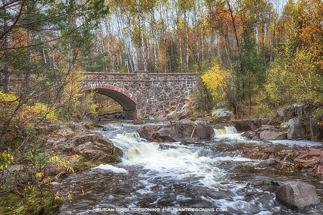 Bridge #7 on Seven Bridges Road, a recently renovated stone arch, spans a rocky stream lined with autumn foliage. Water rushes beneath the bridge, surrounded by trees in shades of yellow, orange, and brown.