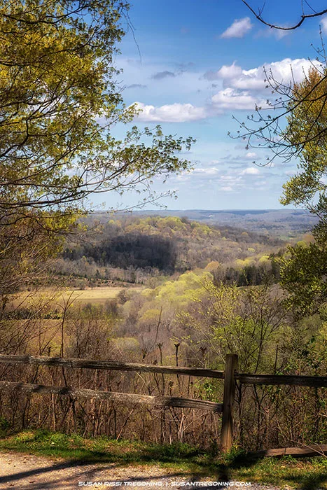 A partial view of the valley seen through a break in the trees along Old Trace Drive, with a rustic wooden fence in the foreground and rolling spring hills under a bright blue sky with scattered clouds.