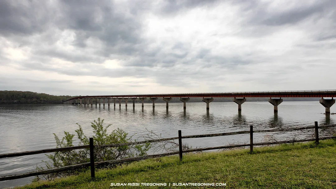   
A long concrete bridge supported by evenly spaced pillars stretches across a calm body of water under an overcast sky. A grassy area with a wooden fence and small shrubs appears in the foreground, with a tree‑covered shoreline in the distance.