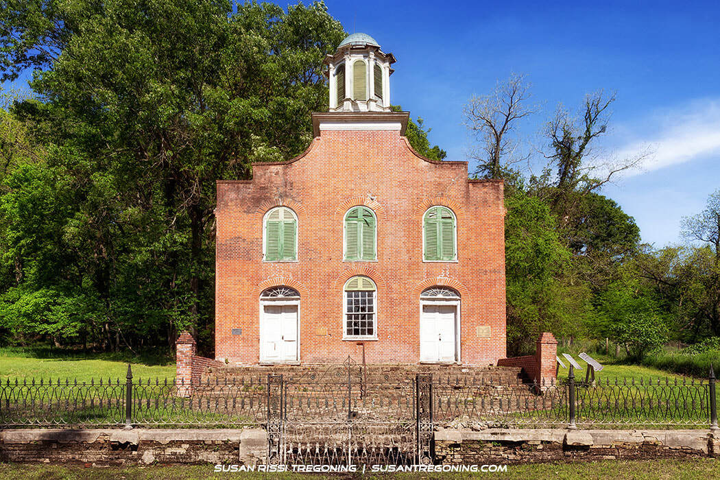 A historic brick building with a curved gable, two arched doorways, and three arched windows with green shutters stands behind a wrought‑iron fence, surrounded by lush green trees and grass in bright daylight.