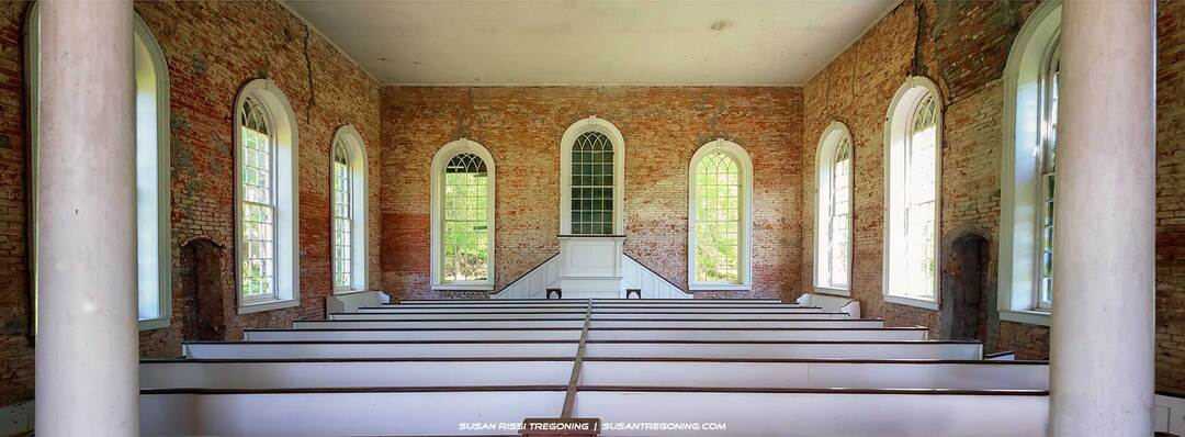 The interior of a historic brick church with tall arched windows, rows of white wooden pews, and a raised central pulpit framed by three large arched windows, all lit by natural light and showing signs of age in the exposed brick walls.