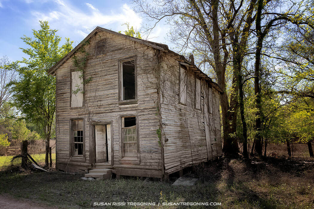 An abandoned two‑story wooden building with peeling paint and vines growing up the walls stands in a grassy clearing beside a dirt path, its windows broken or missing and the front door open, surrounded by tall green trees in bright daylight.