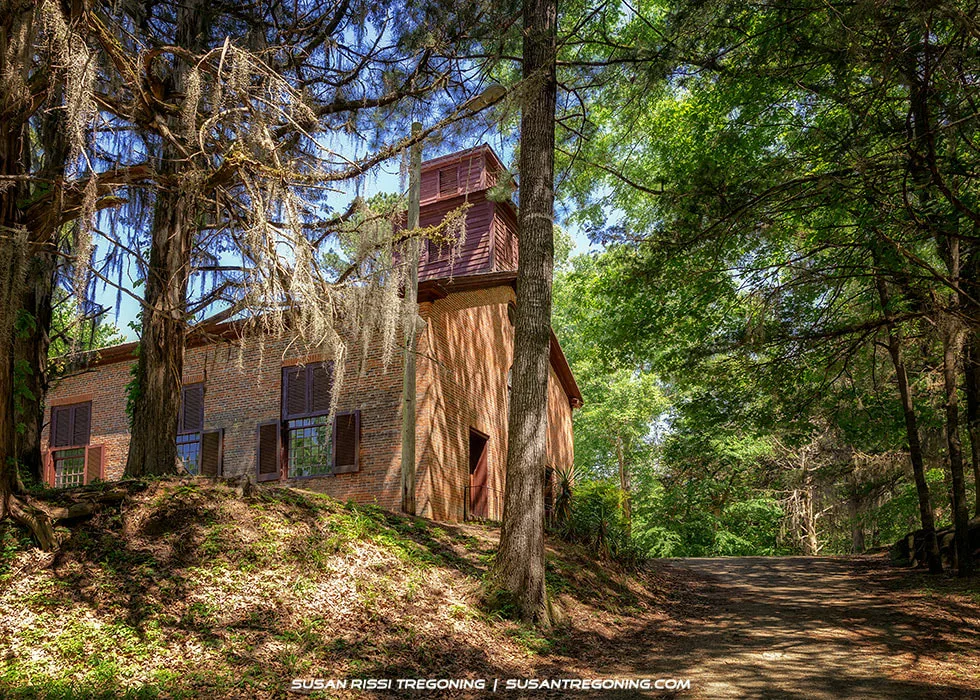 A rustic brick church stands among tall trees in a wooded area, with sunlight filtering through the foliage. The building has large shuttered windows and a wooden tower structure on the roof. A dirt path leads toward the church, and Spanish moss hangs from nearby branches.