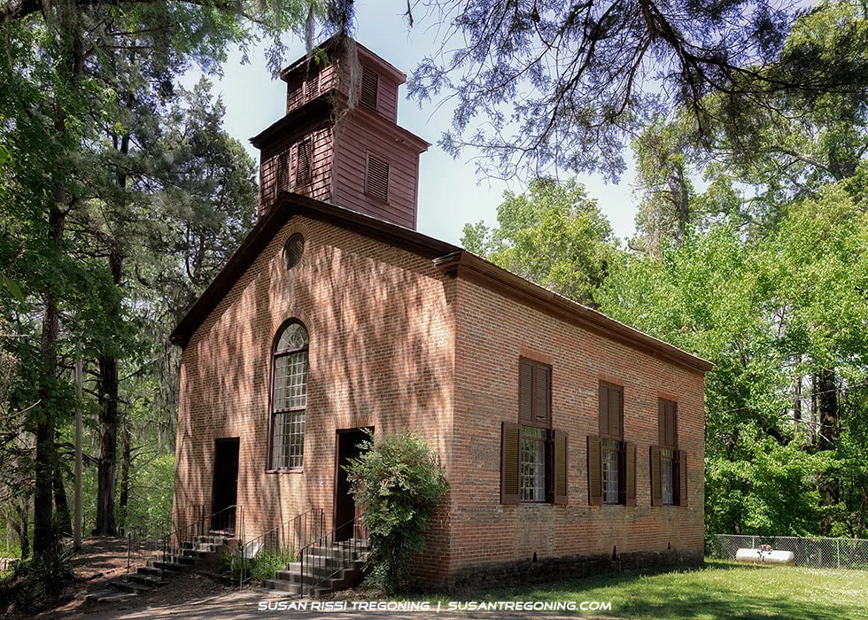 A historic brick church with a steep gabled roof and a square wooden bell tower stands among tall trees. The building has tall arched windows with brown shutters and two staircases leading to separate front entrances. Sunlight filters through the surrounding foliage.