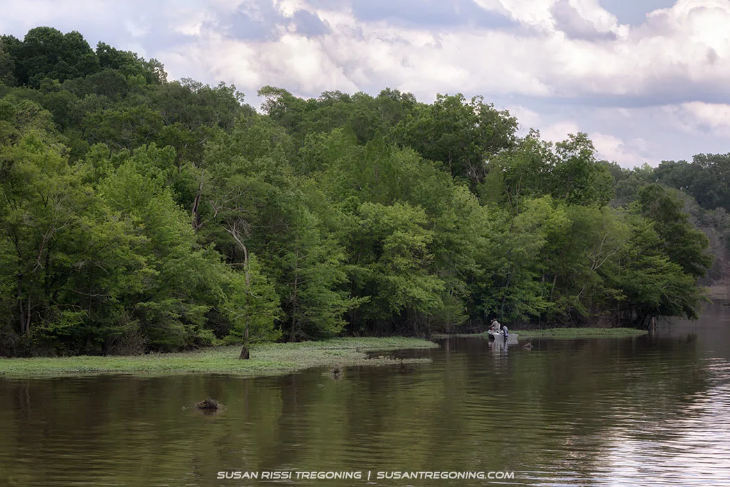 A calm river bordered by dense green forest, with reflections of the trees and cloudy sky on the water. Near the right side of the scene, a small boat with two people sits close to the shoreline.