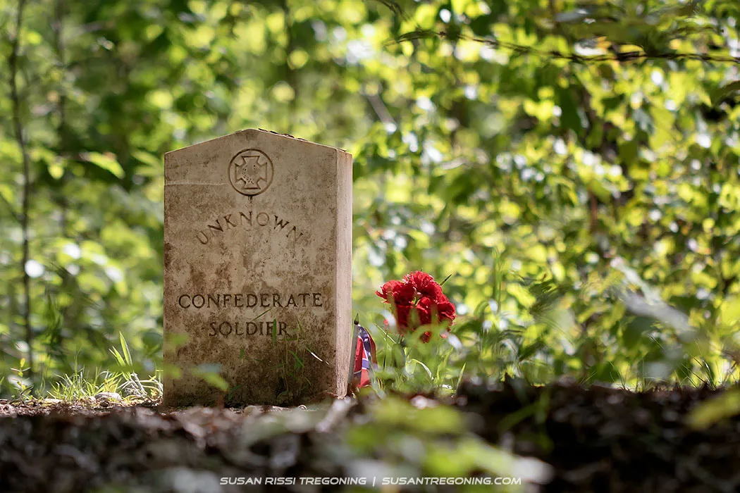   
A single light‑colored gravestone marked “Unknown Confederate Soldier” stands in a wooded area. A carved emblem appears above the inscription, and red flowers are placed at the base of the stone. Sunlight filters through the surrounding green trees, casting soft light across the grave and forest floor.