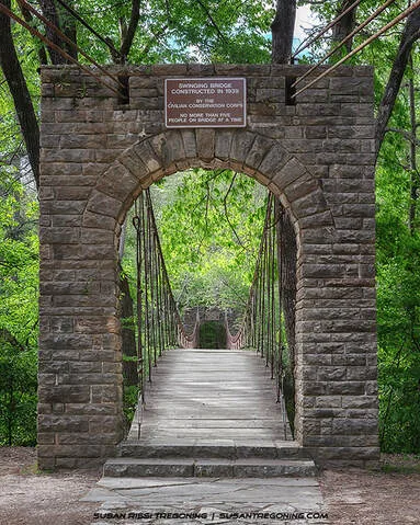 A 1939 stone‑arch entrance frames the wooden‑plank Swinging Bridge at Tishomingo State Park. Steel suspension cables extend into a dense green forest as the bridge spans about 200 feet across Bear Creek. A bronze plaque on the arch identifies it as a Civilian Conservation Corps project.