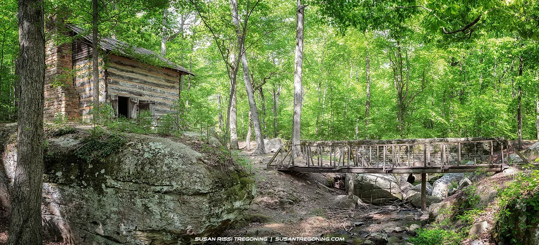 A small 1930s log well house sits atop a mossy rock outcrop in a dense forest at Tishomingo State Park. A narrow wooden footbridge crosses a shallow rocky stream beside it, with sunlight filtering through tall trees and illuminating the rustic cabin and surrounding greenery.