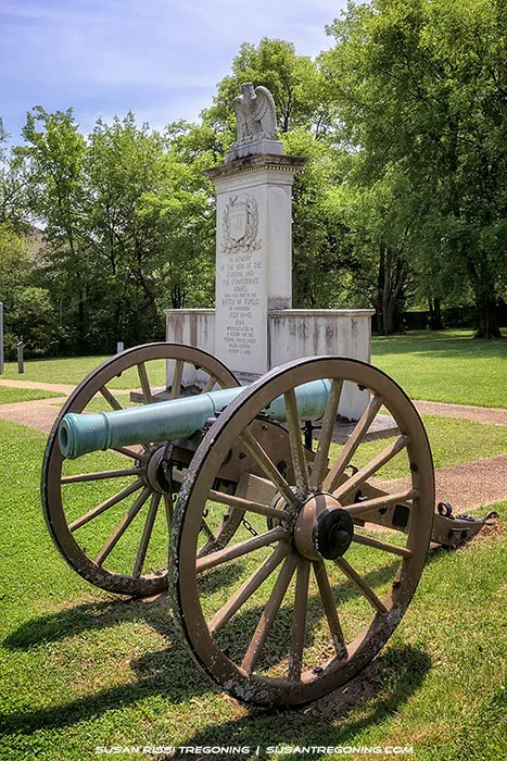 A historic cannon with wooden wheels stands in front of a tall stone monument topped with an eagle at Tupelo National Battlefield. The monument rises from a grassy area surrounded by trees, with a paved walkway leading toward it. Sunlight filters through the foliage, illuminating the cannon and memorial.
