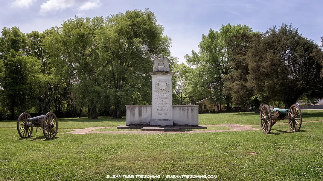 A tall stone monument topped with an eagle stands in a grassy park surrounded by trees. An old cannon on wooden wheels is positioned on each side of the monument. Sunlight filters through the partly cloudy sky, illuminating the memorial and the surrounding green landscape.