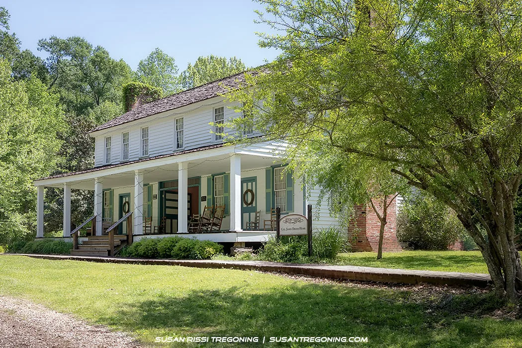 A two‑story white wooden house with a wide front porch and several rocking chairs stands surrounded by green trees and shrubs. Two front doors display decorative wreaths, and a brick chimney rises on the right side of the house. A small sign in the yard identifies the building as the Colonel James Drane House
