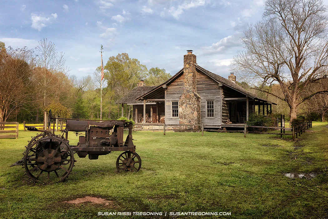 An old rusty tractor with metal wheels sits in front of a log cabin with a stone chimney and a small porch. Rocking chairs are arranged on the porch, and a wooden fence runs along the side of the yard. Trees and grass surround the cabin, and an American flag is visible in the background.