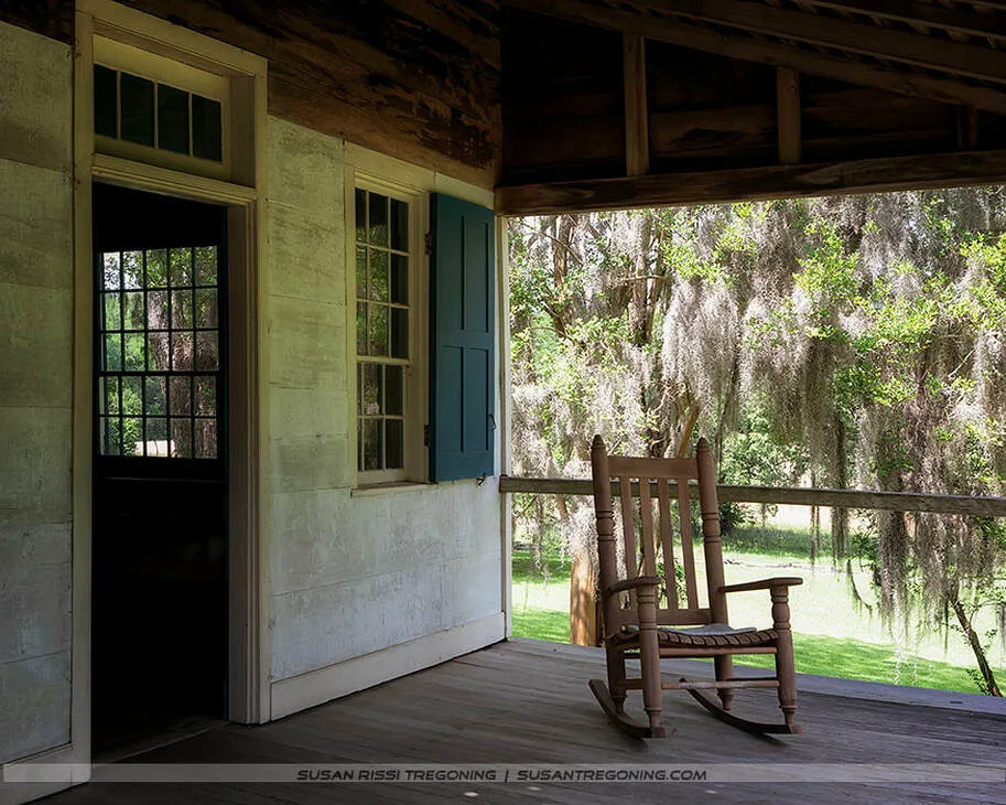 An old wooden rocking chair sits on the shaded front porch of Mount Locust, facing the Natchez Trace. Weathered porch boards and soft afternoon light highlight the age of the historic home, while Spanish moss hangs from a tree in the background.