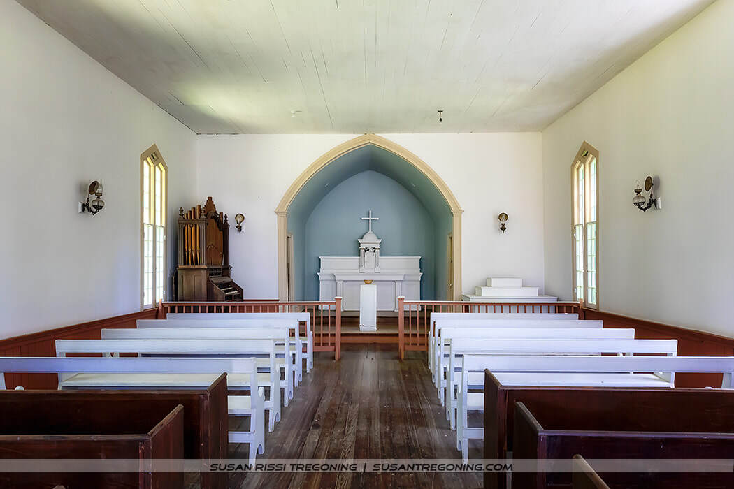 The interior of a small white chapel with wooden pews facing a simple altar set within a blue arched recess. A white cross and pulpit stand at the center, with a pipe organ to the left and tall arched windows bringing in natural light.