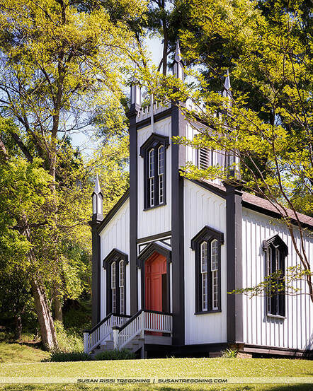 A small white Carpenter Gothic church with pointed arched windows, decorative trim, and a red front door stands surrounded by lush green trees and grass. A short staircase leads to the entrance, and the building appears well maintained in a peaceful, wooded setting.