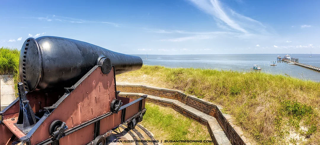 A large historic cannon on a wooden and metal carriage sits on a brick fortification overlooking a body of water. A pier with boats extends into the water in the background, with grassy earthworks and a partly cloudy blue sky completing the scene.