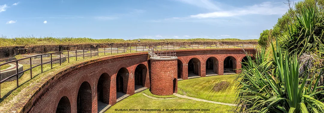   
A panoramic view of a historic brick fort shows a circular courtyard surrounded by arched openings and a central cylindrical tower. Grass covers parts of the interior and the top of the ramparts, with metal railings along the upper walkway. The scene is set under a clear blue sky.