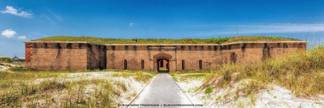 A historic brick fort with a central arched entrance and grass‑covered earthworks stands along a paved walkway surrounded by sand and dune grass. The scene is set against a clear blue sky.