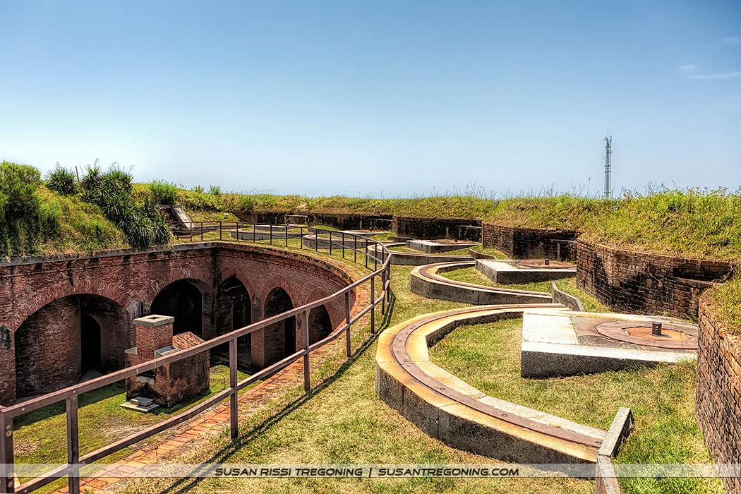 A historic brick fort with circular gun emplacements and arched openings curves along a grassy earthwork, bordered by a metal railing. A tall tower stands in the distant background beneath a clear blue sky.
