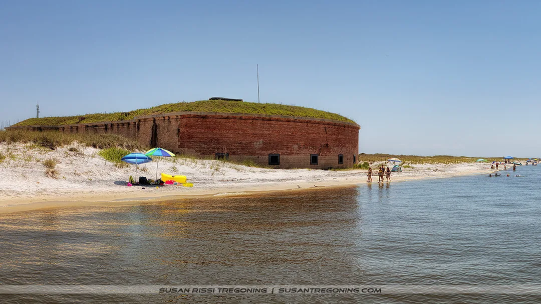 A historic brick fort sits along a sandy beach with people walking, swimming, and sitting under colorful umbrellas near the shoreline. The fort’s red brick walls rise above the sand, topped with grass, beneath a clear blue sky.