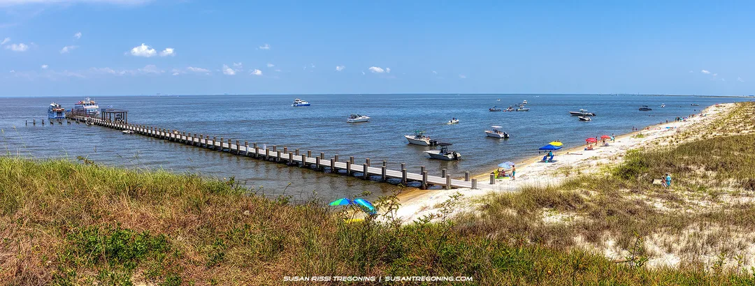A long wooden pier extends over calm blue water with several anchored boats nearby. People sit under colorful umbrellas and relax along the sandy beach, with grassy dunes in the foreground and a clear sky above.