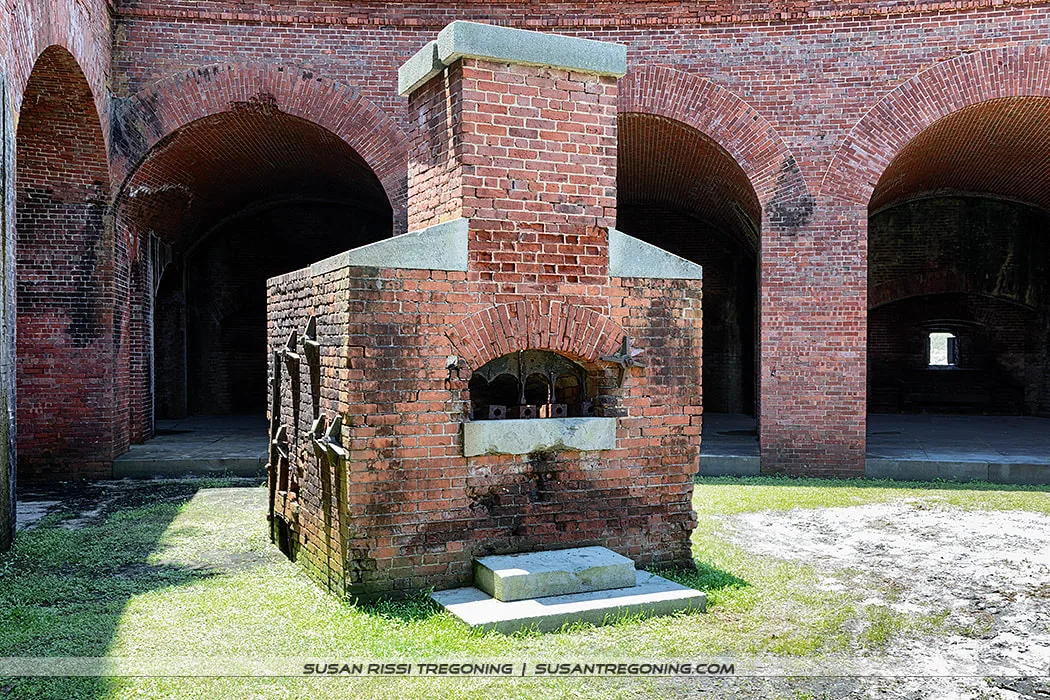A brick Hot Shot Furnace with an arched firebox opening and a tall chimney stands inside a historic fort. The structure sits on a grassy floor beneath high brick arches, illuminated by natural light.
