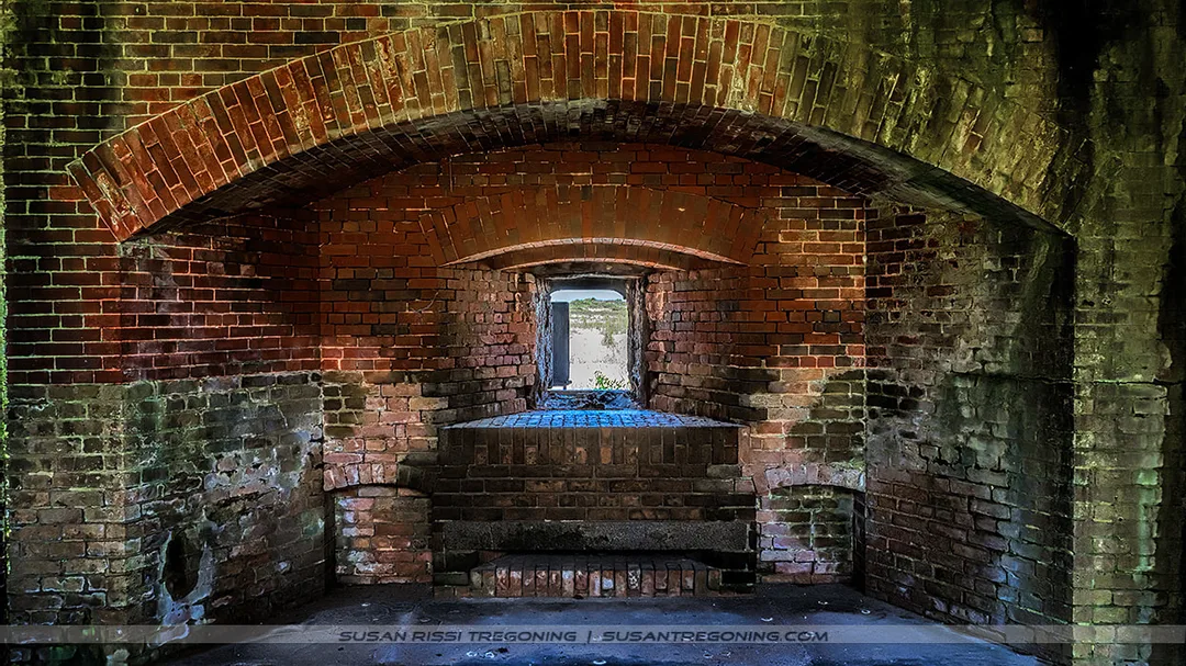 A brick casemate inside a historic fort features a series of arched ceilings and walls leading toward a bright rectangular opening at the far end. Natural light highlights the weathered brick and the shadowed interior passage.