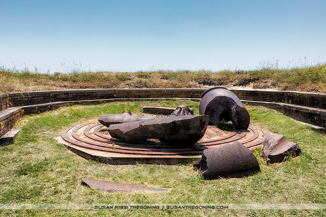 Broken pieces of a large historic cannon rest inside a circular brick and concrete gun emplacement with concentric metal mounting rings. Grass fills the interior area, and a low brick wall curves around the structure beneath a clear blue sky.