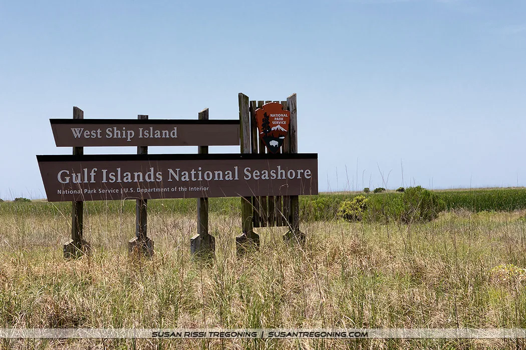 A wooden sign for West Ship Island stands in a grassy coastal area. The sign reads “West Ship Island, Gulf Islands National Seashore” with the National Park Service emblem on the right, set against vegetation and a clear blue sky.