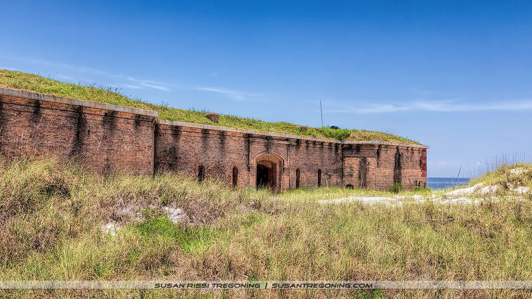 A historic brick fort with arched openings and grass-covered earthworks stands near a sandy area with coastal grasses. The ocean is visible behind the structure under a clear blue sky.
