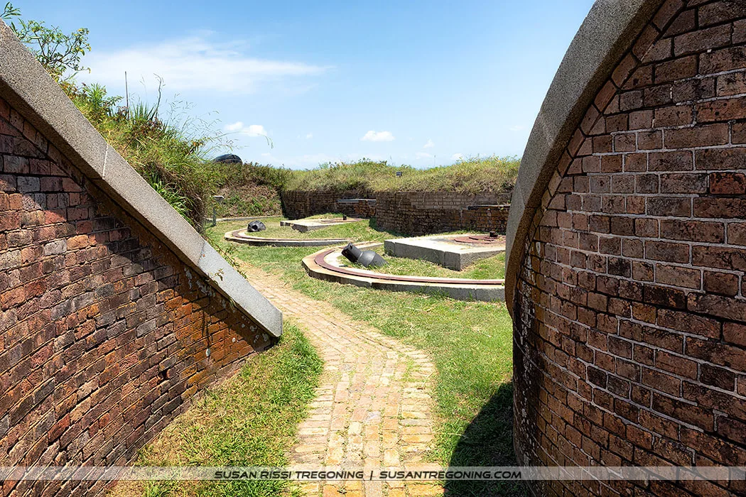   
A curved brick fort wall borders a narrow brick pathway that winds through grassy terrain. In the distance, circular gun mounts with historic cannons sit on a raised earthwork beneath a clear blue sky.
