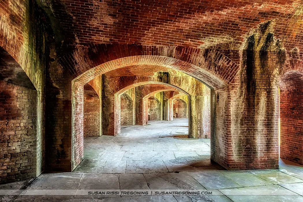 A series of brick archways forms a long corridor inside a historic fort. The repeating arches create a tunnel‑like passage with textured brick walls and a stone floor, illuminated by natural light.