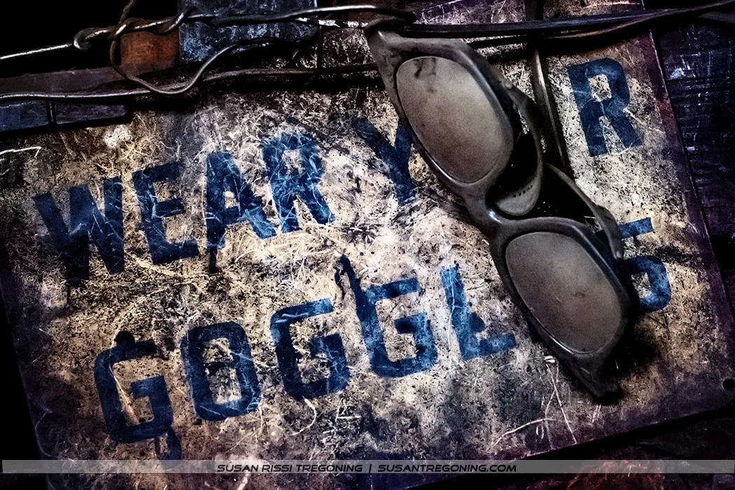 A pair of old, dusty protective goggles rests on a worn metal safety sign with faded blue lettering that reads “WEAR YOUR GOGGLES.” The surface shows scratches, rust, and age, giving the scene a vintage industrial feel. The composition highlights the importance of eye protection in historic workshop environments.