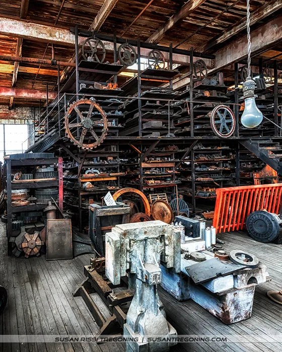 Shelves inside a historic workshop filled with metal patterns and mechanical parts, including gears, pulleys, and wheels stacked in rows. The space features worn wooden floors, aged brick walls, and a single hanging light bulb illuminating the textures of the metal and wood. The scene captures the dense, organized storage typical of an early industrial pattern shop.