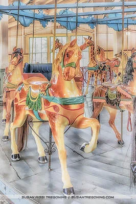 Brightly painted carousel horses stand on a vintage Dentzel carousel, decorated with ornate saddles, bridles, and colorful detailing. The horses are arranged around the central platform beneath a canopy with blue decorative panels, all set inside a wooden pavilion that adds to the historic charm.