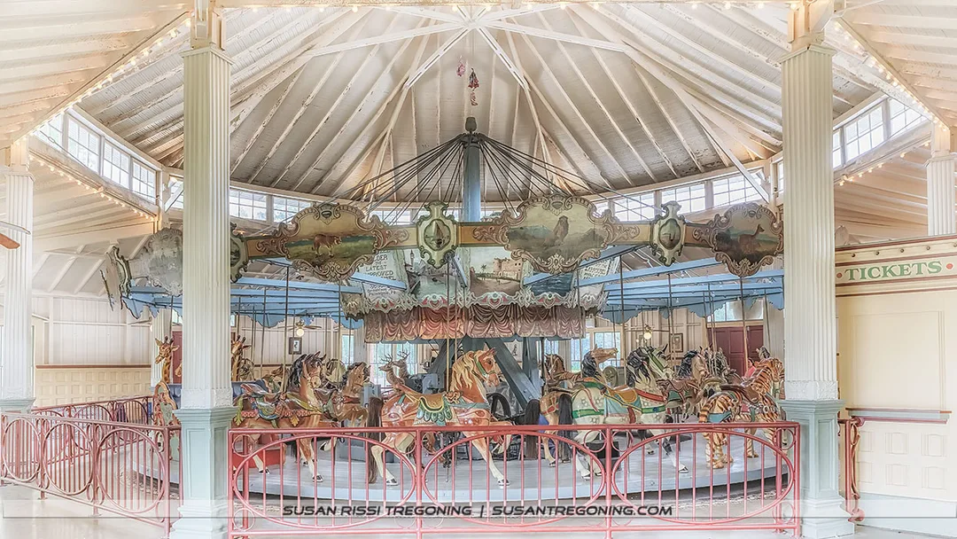 An antique carousel sits inside a round pavilion with a high white wooden ceiling supported by large columns. The carousel features brightly painted horses with ornate saddles and decorative panels above them. A red railing circles the ride, and a small ticket booth stands to the side. Soft, warm light highlights the vintage charm of the historic structure.
