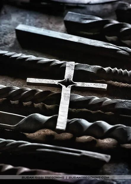 A collection of forged metal pieces arranged on a surface, including a central metal cross with a star‑shaped cutout and several twisted or textured metal rods placed around it. The lighting highlights the metallic surfaces and craftsmanship, giving the scene a handcrafted, blacksmith‑workshop feel.
