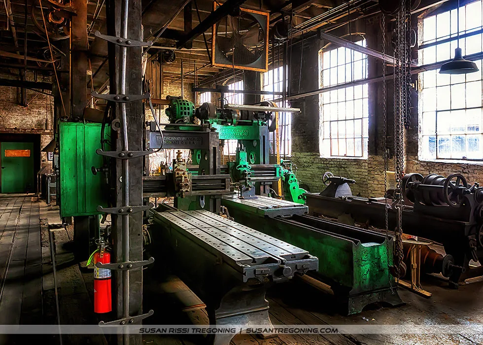 A historic machine shop interior with large metal planers and industrial equipment arranged along a brick‑walled workspace. A green Cincinnati Progress Co. planer sits in the foreground, with its long metal bed, cutting head, and overhead pulley system visible. Sunlight filters through tall multi‑pane windows, illuminating the worn floors, exposed beams, and surrounding early‑20th‑century machinery. A red fire extinguisher is mounted on a nearby column.