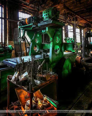 A historic machine shop interior featuring a large green metal planer with a motor mounted on top and a flat cutting bed. In front of the machine sits a metal cart filled with cutting tools and drill bits organized in trays. The workshop has brick walls, tall industrial windows, and warm overhead lighting that highlights the surrounding early‑20th‑century machinery.
