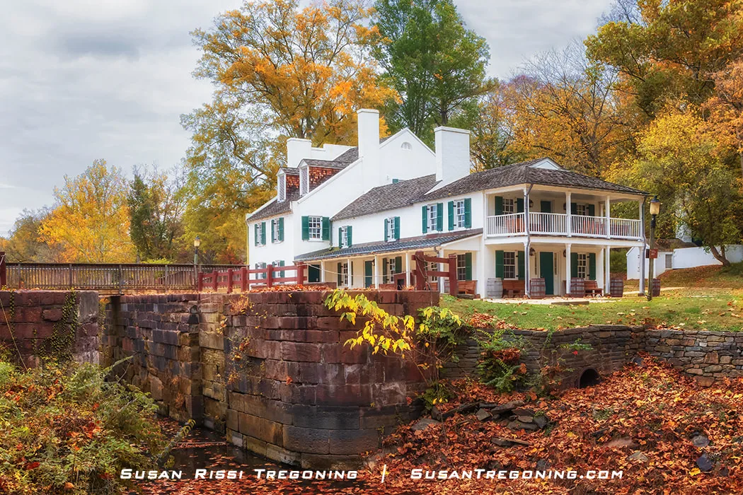 A white historic lockhouse with green shutters and a wraparound porch stands beside a stone canal lock surrounded by vibrant autumn trees in shades of yellow, orange, and green. Still water fills the lock in the foreground under clear fall light.