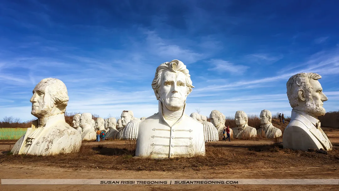 A field filled with large, weathered concrete busts of former U.S. presidents, arranged in rows under a bright blue sky. A prominent bust wearing a military-style jacket stands in the foreground, with additional presidential heads and a few people walking among them in the background.