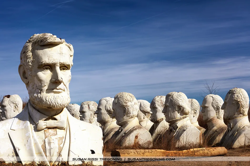 A large, weathered concrete bust of Abraham Lincoln stands in the foreground of an open field, with rows of additional presidential heads behind it. All of the sculptures show visible cracks and discoloration, set against a clear blue sky above dry grass and dirt.