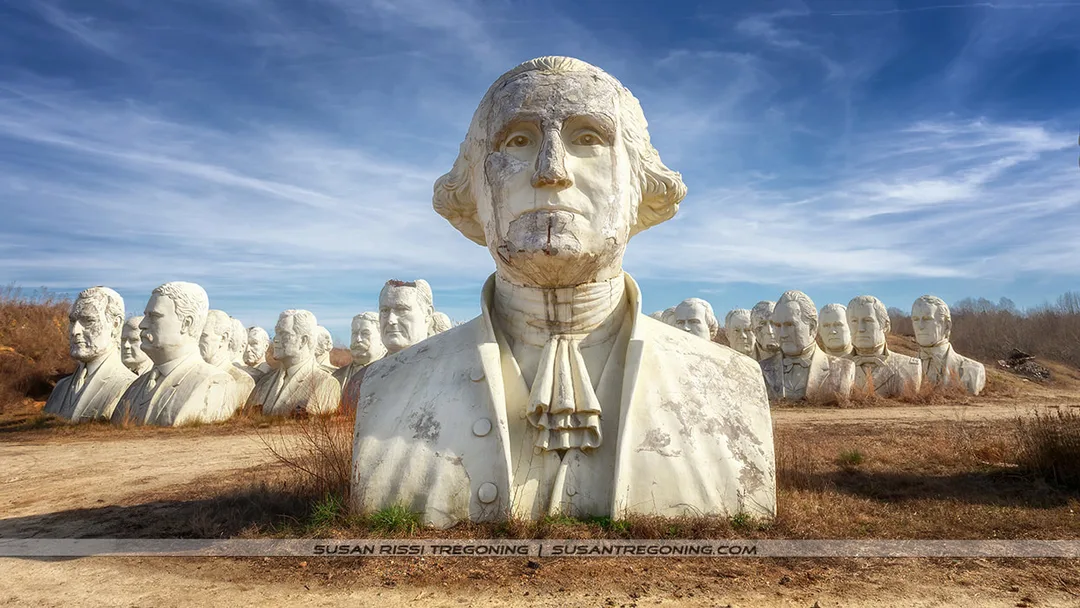 Large, weathered concrete busts of former U.S. presidents stand in an open field, with George Washington’s cracked and discolored head in the foreground. Additional presidential heads fill the background under a partly cloudy blue sky, surrounded by dry grass and sparse vegetation.