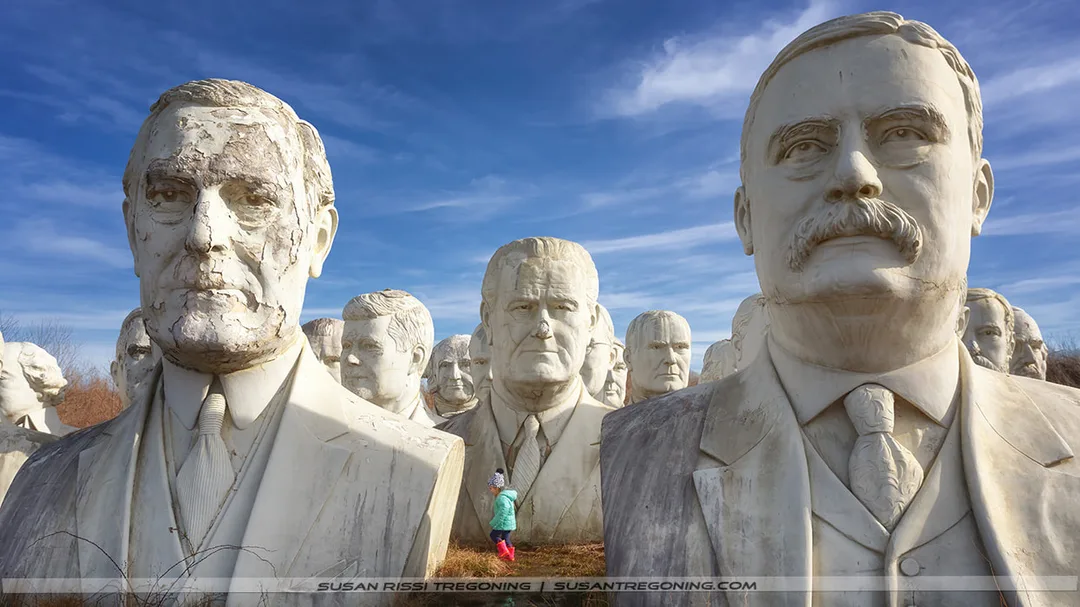 Large, weathered concrete busts of Theodore Roosevelt, Woodrow Wilson, and Lyndon Johnson stand outdoors in a grassy field. A child in a turquoise jacket and pink pants stands among the massive sculptures, emphasizing their scale. The statues show surface cracks, discoloration, and erosion beneath a sky with thin, wispy clouds.
