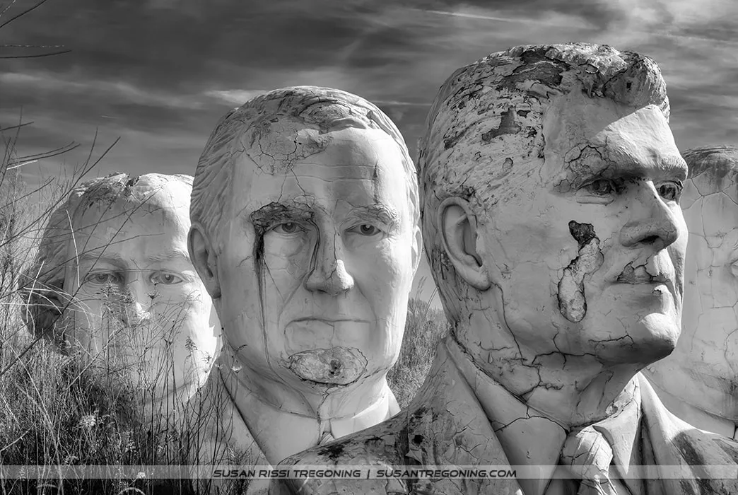 Large, weathered concrete busts of Ronald Reagan, Warren G. Harding, and John Adams stand outdoors in a grassy field. The black‑and‑white image highlights the cracked surfaces and erosion on the sculptures. John Adams’ bust is partially obscured as it peeks through tall weeds in the foreground, with a partly cloudy sky behind them.