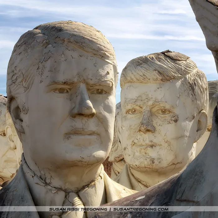 Large, weathered concrete busts of Jimmy Carter and John F. Kennedy stand outdoors in a field. Their surfaces show cracks, discoloration, and erosion from long exposure to the elements. Additional presidential busts appear in the background beneath a clear blue sky.