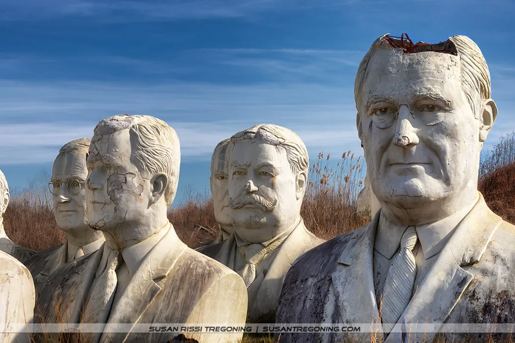 A group of large, weathered concrete presidential busts stands outdoors in a field. Visible are Franklin D. Roosevelt, George W. Bush, William Howard Taft, Harry S. Truman, and William McKinley. FDR’s bust has the entire top of his head missing, exposing internal metal reinforcement. The surrounding sculptures show cracks, discoloration, and erosion beneath a clear blue sky.
